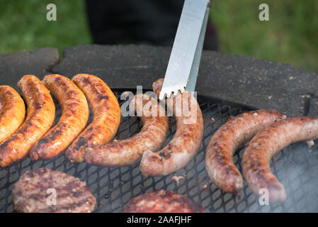 Steel tongs grabbing a sausage being cooked over an open grill Stock Photo