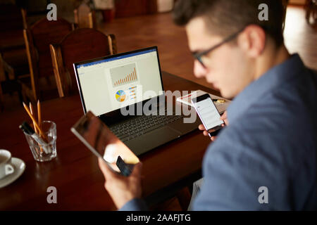 Attractive man in glasses working with multiple electronic internet devices. Freelancer businessman has laptop and smartphone in hands and laptop on Stock Photo