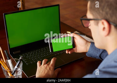 Back view of businessman using smartphone and laptop typing in office. Green screen mockup. Freelancer workspace. People and technology. Copyspace and Stock Photo