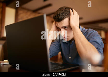 Feeling tired. Frustrated young handsome man looking exhausted and sick while sitting at his working place with laptop on the table. Freelancer has Stock Photo