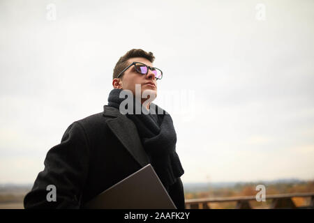 Handsome attractive businessman using a laptop on office terrace. Stylish man in glasses coat and scarf standing on balcony with laptop. Smart casual Stock Photo