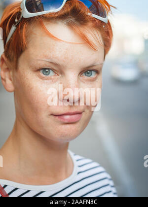 Close-up portrait of a middle-aged red-haired woman looking at the camera and smiling on a blurred street background, selective focus Stock Photo