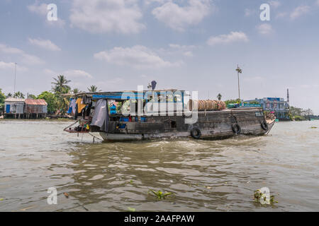 Cai Be, Mekong Delta, Vietnam - March 13, 2019: Along Kinh 28 canal. Trading family old wooden barge sails away towards Mekong River under blue clouds Stock Photo