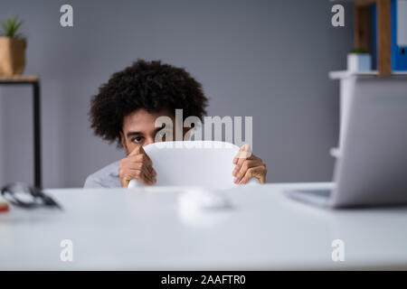 Frightened Young Businessman Hiding Behind Chair At Workplace Stock Photo