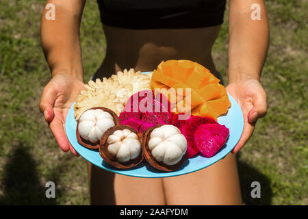 A girl in the green jungle is holding a plate with freshly cut and peeled tropical fruits. Stock Photo