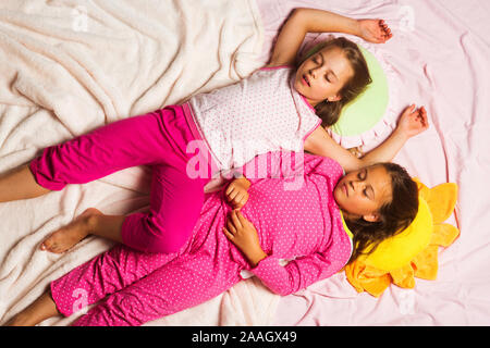 Children with sleepy faces lie close on light pink blanket background. Kids in pink pajamas sleep with put up hands. Schoolgirls have pajama party with funny pillows. Childhood and happiness concept. Stock Photo