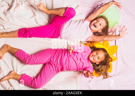 Children with smiling faces lie on light pink blanket background and hold hands up. Schoolgirls have pajama party with funny pillows. Kids in pink pajamas, top view. Childhood and happiness concept. Stock Photo