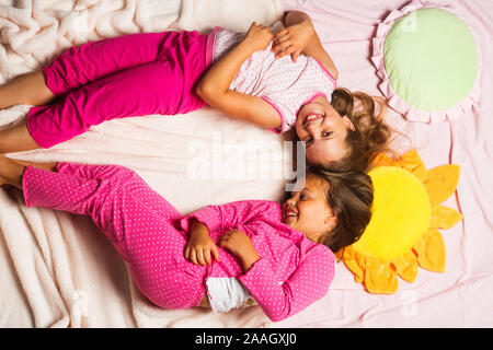 Children with happy faces lie close on light pink blanket background. Kids in pink pajamas put heads together. Schoolgirls having pajama party with funny pillows. Childhood and happiness concept. Stock Photo