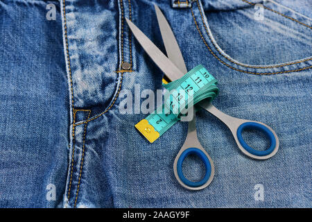 Making clothes and design concept. Metal scissors wound around measure tape on jeans. Jeans crotch, pocket and belt loops, close up. Tailors tools on denim fabric, selective focus. Stock Photo
