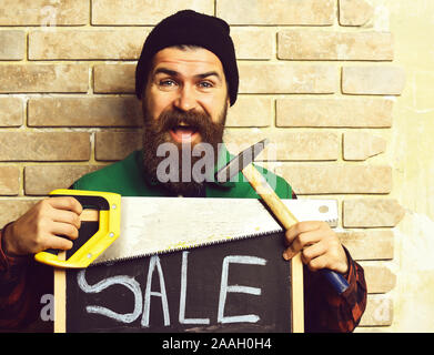 bearded foreman, long beard, brutal caucasian hipster with moustache holding various building tools: saw, hammer and board with inscription sale, happy smiling face, brick wall studio background Stock Photo