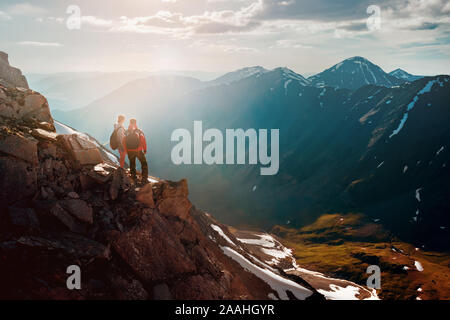 Two hikers male and female stands on a cliff in big mountains and enjoys sunset Stock Photo