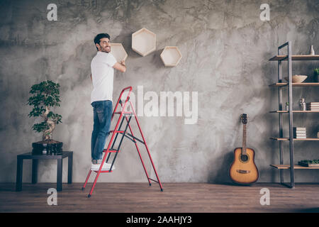 Full length profile view photo of mixed race guy installing wall shelf standing stepladder repair of new flat handmade working with pleasure hanging Stock Photo