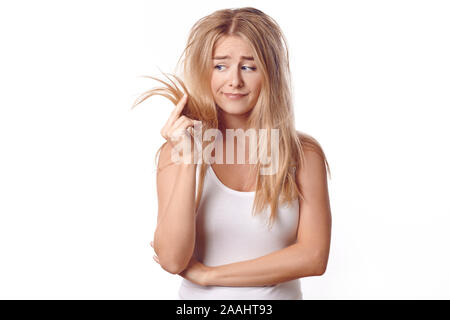 Pretty young woman having a bad hair day standing looking dejectedly at the dry ends of her tousled long blond hair as she holds them up in her hands Stock Photo