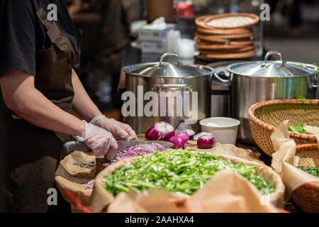 Cook's hands in cellophane gloves cutting red onion into thin slices. Cooking vegetable ingredients. Against the background of lettuce leaves and pans Stock Photo