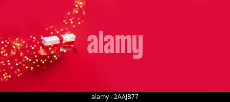 Big gift box wrapped in wrapping pink paper red satin ribbon bow, with shiny glitter flying in the air on a red background with copy space. Stock Photo