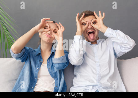 Relationnship Concept. Young couple sitting on sofa studio isolated on grey making eyeglasses with hands grimacing playful Stock Photo