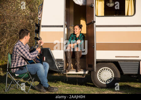 Girlfriend sitting on the stairs of retro camper van and listening her boyfriend playing on guitar. Stock Photo