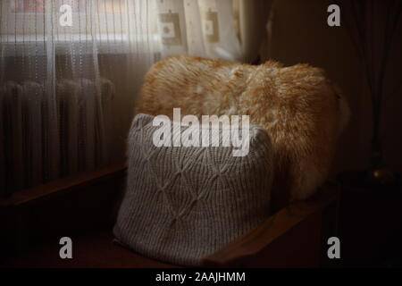 Knitted pillow and fur cape on a chair in the living room by the window. Stock Photo