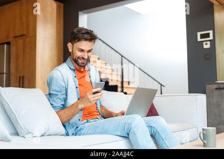 Young adult man sitting at home, using smartphone and laptop Stock Photo