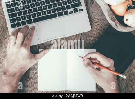 top view of man taking notes on note pad while working on laptop computer on wooden table Stock Photo