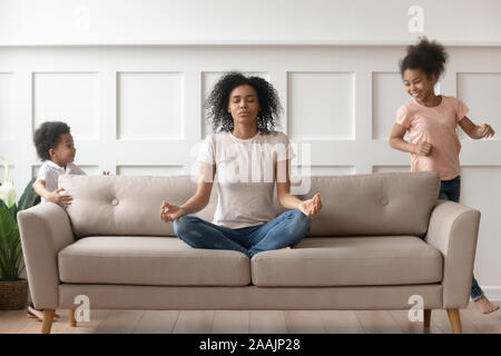 While noisy kids running young african mother meditates on couch Stock Photo