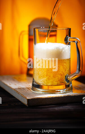 Pour Light Beer into Mugs. Light Beer Glass Mugs with White Foam on Woowen Table and Yellow Background Stock Photo