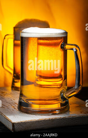 Two Light Beer Mugs with White Foam on Dark Wooden Table and Yellow Background Stock Photo