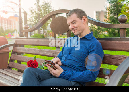 Happy man in a blue suit and with a red rose, sits on a park bench and carries on correspondence by phone in the bright rays of the sun Stock Photo