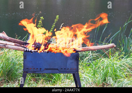 Firewood burns in grill, preparing coals for grilling. Barbecue on nature. Wood fire prepared for BBQ. Grilling season near lake or river. Stock Photo