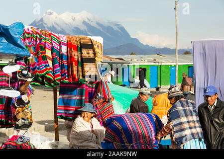 An indigenous woman selling traditional colourful Bolivian fabric at a street market in El Alto, Bolivia, with the peak of Illimani in the background. Stock Photo