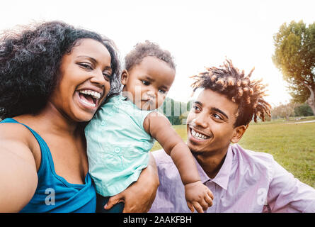 Happy African family doing selfie photo with mobile phone in a public park outdoor - Mother and father having fun with their daughter during a weekend