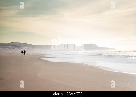 Silhouettes of people walking along Guincho beach - Praia do Guincho - on hazy windy and sunny day in winter with waves rolling in Portugal Stock Photo