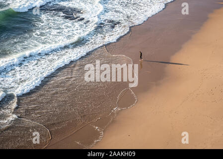 Waves rolling in to |Atlantic coast with empty beach with single barefoot walker casting long shadow on sand in Azenhas do Mar in Portugal Stock Photo