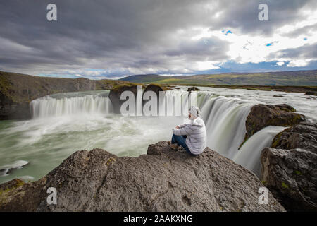 Man sitting on edge of rock, Godafoss waterfall, Baroardalur, Iceland Stock Photo