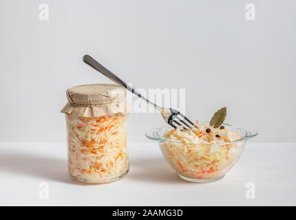 Homemade sauerkraut. Fermented food. Sauerkraut with carrots in glass jar and bowl with fork on wooden white background. Stock Photo