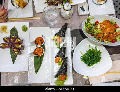 Japanese flat lay of delicious dishes. Black Uramaki of salmon, Temaki rice cones, fresh salmon tartar, seaweed with soy sauce, surimi salad and water Stock Photo