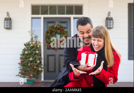 Young Mixed Race Couple Exchanging Gift On Front Porch of House with Christmas Decorations. Stock Photo