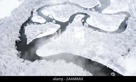 A drone / aerial view of a windy river, surrounded by snow covered trees, with clouds floating over the top. Stock Photo