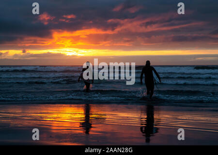 Surfers walking towards the Ocean on the sandy beach during a dramatic summer sunset. Taken in Seaside, Oregon, United States of America. Stock Photo