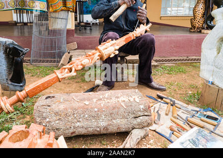 black African man chisels and carves a piece of wood or wooden pole with traditional figurines outdoors in Graskop, Mpumalanga, South Africa Stock Photo