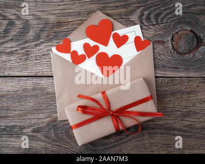 card and red hearts in open envelope from brown Kraft paper. Gift box with red ribbon on wooden aged vintage background. Greetings with Valentine's da Stock Photo