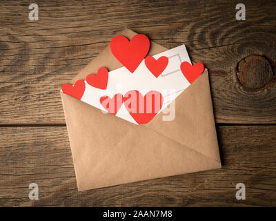card and red hearts in open envelope from brown Kraft paper on wooden aged vintage background. Greetings with Valentine's day Stock Photo