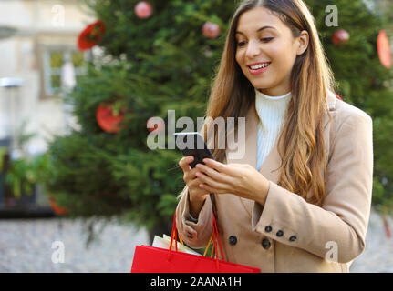 Christmas woman buying online on the smart phone in the street with christmas tree on background. Stock Photo
