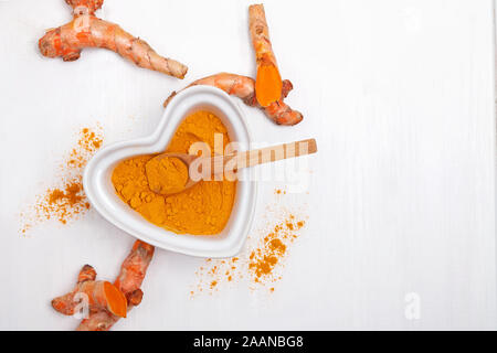 Ground turmeric in heart ahaped bowl and roots on white table top view Stock Photo