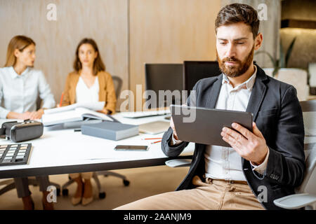Confident businessman working on the digital tablet while sitting with colleagues at the working place of some creative office studio Stock Photo
