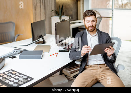 Confident businessman working on the digital tablet while sitting at the working place of some creative office studio Stock Photo