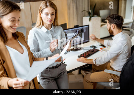Creative office employees working on some architectural project, holding blueprints and workign on the computer in the modern office studio Stock Photo