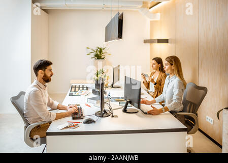 Young employees dressed casually doing some creative work at the large table with computers in the office. Concept of an architectural business and creative work Stock Photo