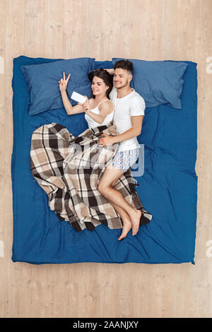 Bedtime. Young couple lying on bed under blanket top view hugging taking selfie on phone cheerful Stock Photo