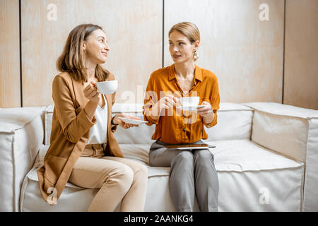 Two female colleagues having cheerful conversation, sitting together on the couch during a coffee break in the office Stock Photo